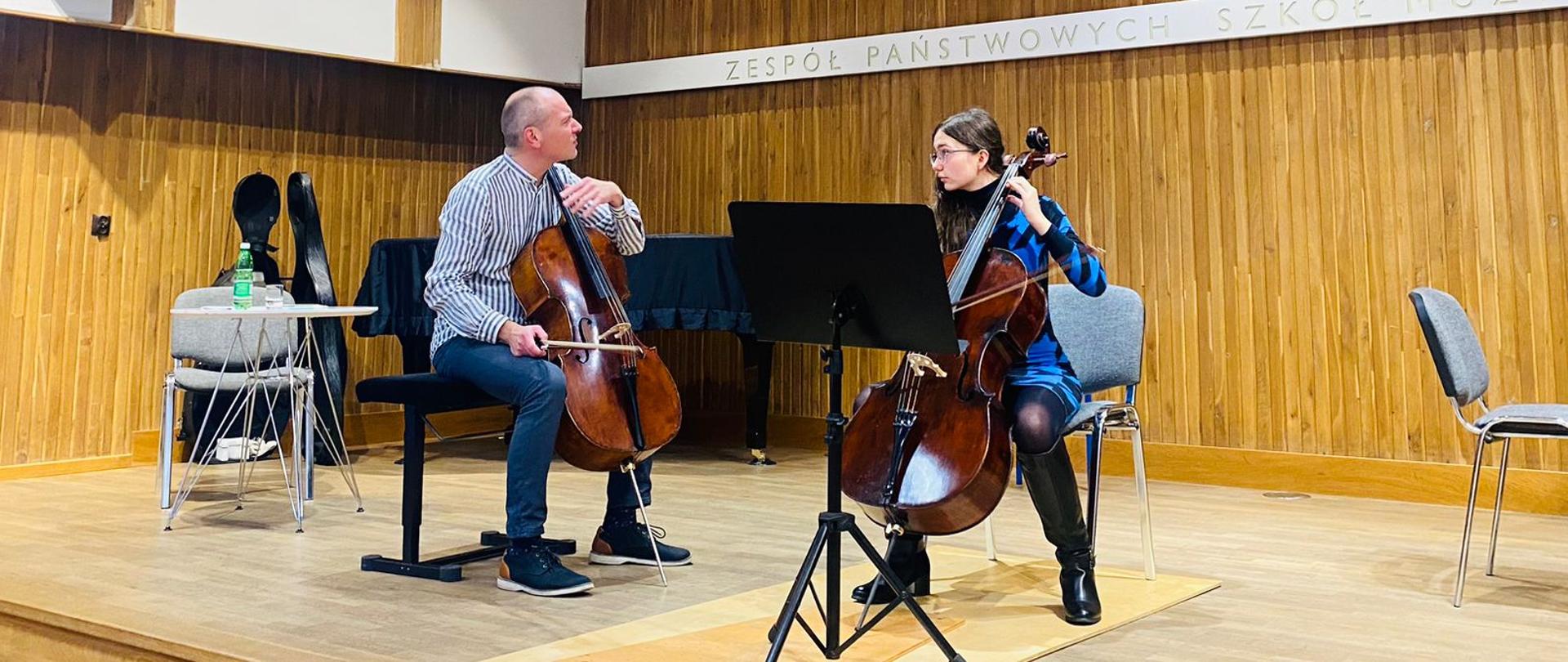 Zdjęcie przedstawia: Wiolonczelista i wiolonczelistka na scenie z jasnego drewna, przed uczennicą stoi pulpit na nuty, po prawej stronie widać fragment fortepianu po lewej stronie cały fortepian w tle za uczennicą.