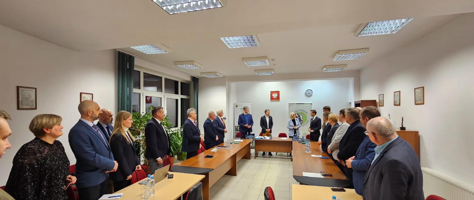 Uczestnicy spotkania stoją za stołami konferencyjnymi ustawionymi w literę C