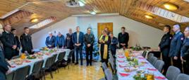 Zdjęcie dokumentujące oficjalną część uroczystości. W centrum, obok choinki, stoi ksiądz ubrany w czarną sutannę i złotą stułę, czytający z małej książeczki (modlitewnika). Zebrani wokół strażacy w mundurach oraz goście cywilni stoją w milczeniu z pochylonymi głowami. Światło wpadające przez okna dachowe oraz ciepłe oświetlenie sufitowe podkreślają domową, a jednocześnie podniosłą atmosferę spotkania w drewnianym wnętrzu.