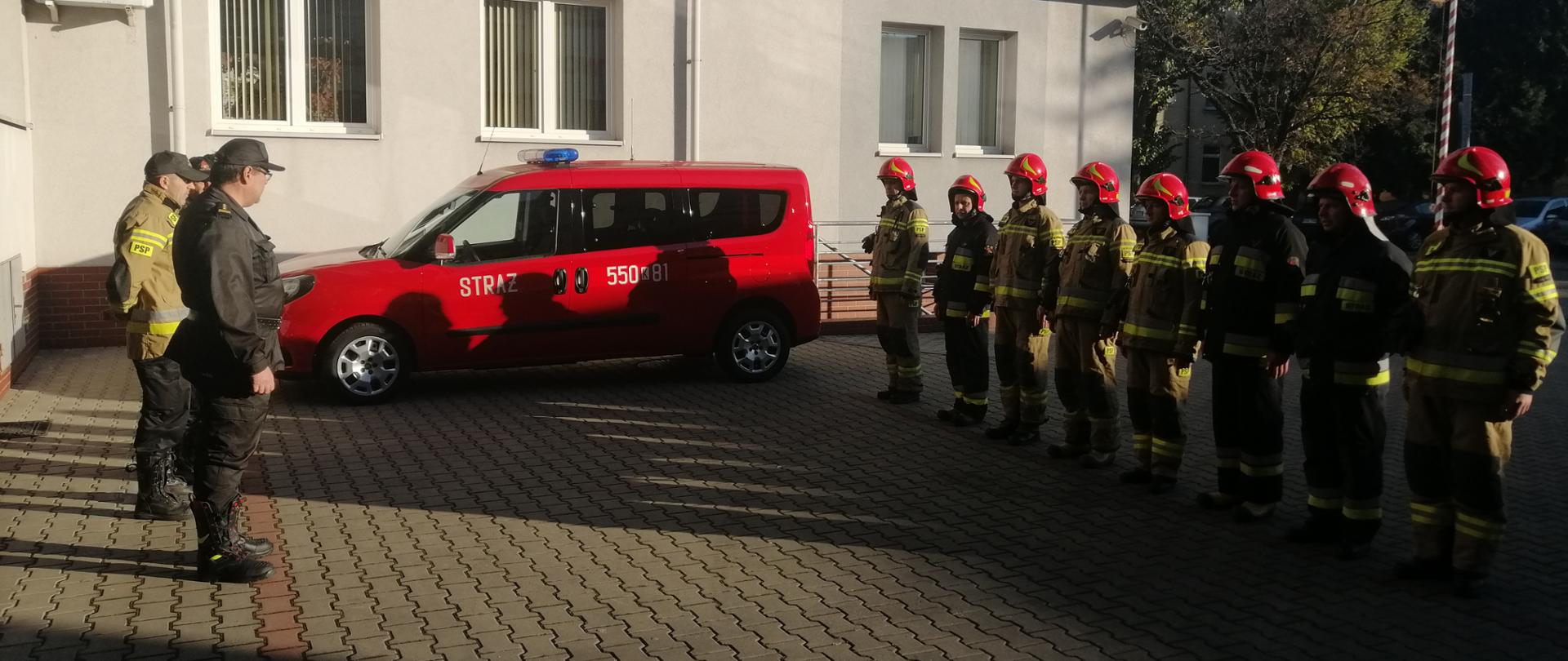 Plac przed jednostką straży pożarnej. Po prawej strażacy w pełnym umundurowaniu bojowym ustawieni w szeregu. Po lewej Komendant Powiatowy PSP w Wadowicach. Obok niego w szeregu komisja przeprowadzająca przegląd.
