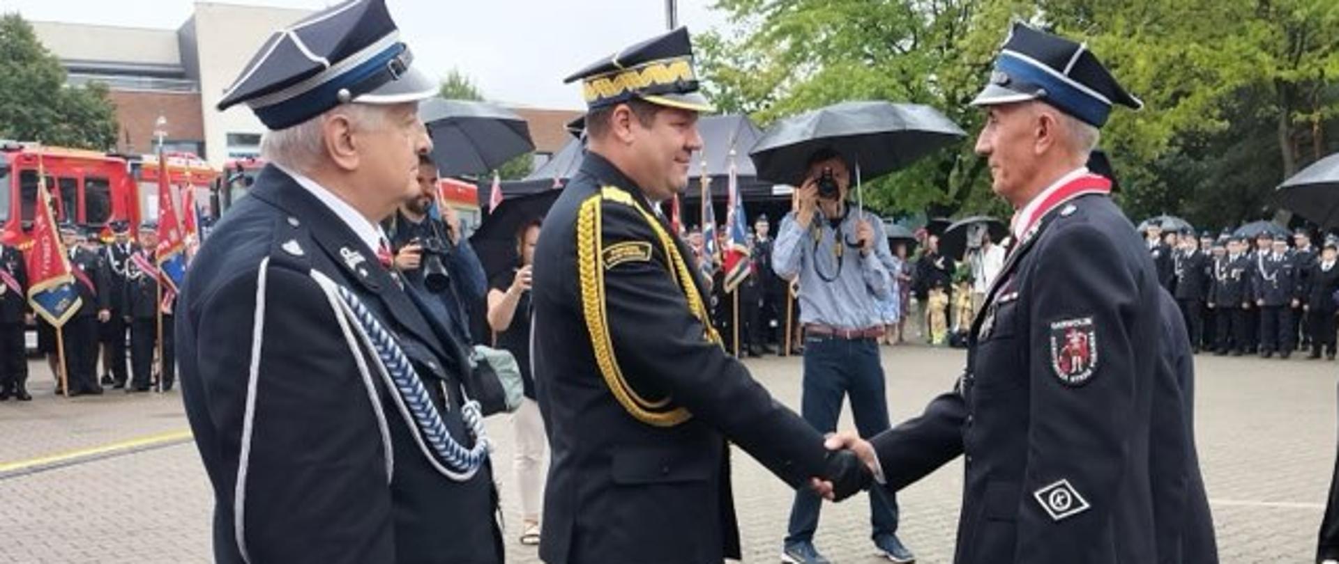  Dwóch strażaków w mundurach stojąc na przeciwko siebie uściskuje sobie prawe dłonie. Obok nich stoi inny strażak w mundurze. W tle widoczne są samochody strażackie, sztandary i zgromadzeniu ludzie.