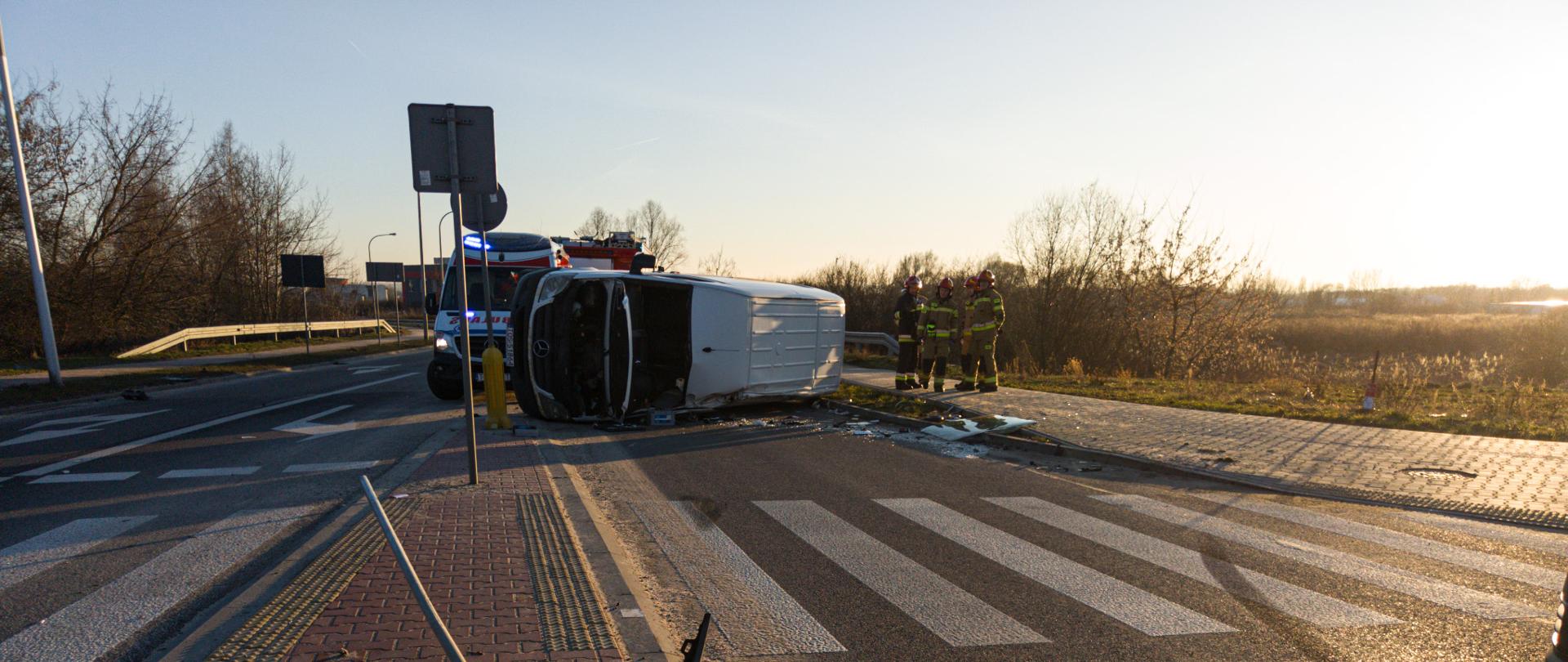 Na pierwszym planie rozbity zderzak przy przejściu dla pieszych. Na dalszy planie wywrócony na bok samochód koloru białego. Przy rozbitym aucie stoi 3 strażaków oraz karetka pogotowia ratunkowego. Pora dzienna, słoneczna.