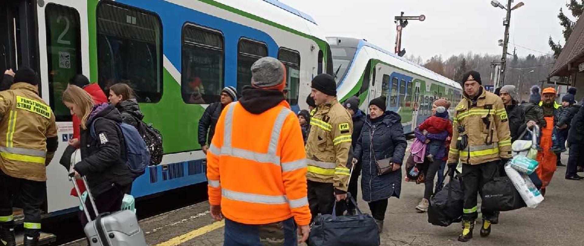 Zdjęcie przedstawia strażaków pomagających w przejściu oraz przeniesieniu rzeczy uchodźców z Ukrainy do podstawionego składu wagonów.
