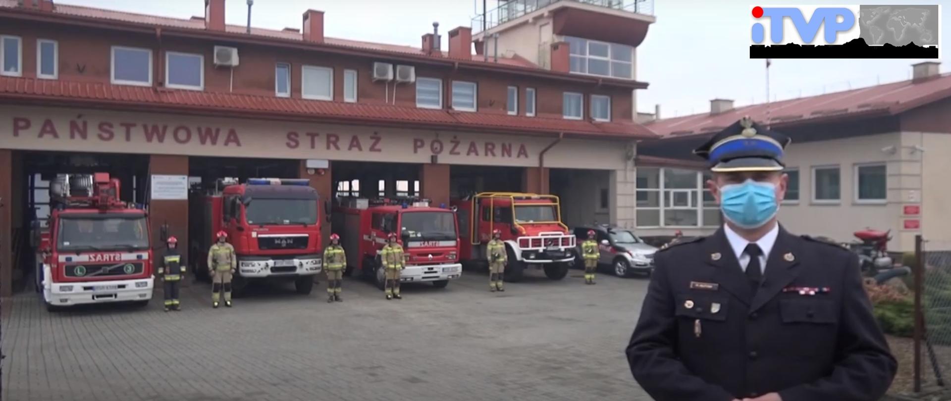 Na zdjęciu po prawej stronie stoi w umundurowaniu wyjściowym brygadier Rafał Rutyna Komendant Powiatowy Państwowej Straży Pożarnej w Strzyżowie. W tle widoczny budynek Komendy Powiatowej oraz garaże jednostki ratowniczo-gaśniczej. Bramy otwarte, samochody stoją w połowie na placu zewnętrznym. Od lewej strony stoi samochód specjalny z podnośnikiem, następnie 3 samochody gaśnicze oraz samochód operacyjny. Przed samochodami stoją strażacy w umundurowaniu specjalnym. Nad bramami widoczny napis Państwowa Straż Pożarna. W prawym górnym rogu logo telewizji ITVP.
