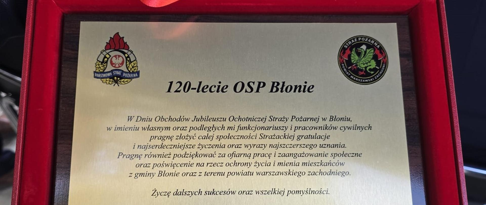 Na zdjęciu widoczna jest pamiątkowa tablica umieszczona w otwartym, czerwonym etui wyściełanym satyną lub aksamitem.
Tablica i Treść
Centralny element to prostokątna tablica (prawdopodobnie metalowa lub z laminatu w kolorze złota/brązu) z grawerowanym (lub drukowanym) tekstem w języku polskim:
1. Nagłówek: 120-lecie OSP Błonie (Ochotnicza Straż Pożarna w Błoniu).
2. Logotypy: Po lewej i prawej stronie u góry widoczne są emblematy:
o Lewy: Państwowej Straży Pożarnej (Państwowa Straż Pożarna).
o Prawy: Państwowej Straży Pożarnej (Państwowa Straż Pożarna).
3. Tekst życzeń i podziękowań:
"W Dniu Obchodów Jubileuszu Ochotniczej Straży Pożarnej w Błoniu, w imieniu własnym oraz podległych mi funkcjonariuszy i pracowników cywilnych pragnę złożyć całej społeczności Strażackiej gratulacje i najserdeczniejsze życzenia oraz wyrazy najszczerszego uznania. Pragnę również podziękować za ofiarną pracę i zaangażowanie społeczne oraz poświęcenie na rzecz ochrony życia i mienia mieszkańców z gminy Błonie oraz z terenu powiatu warszawskiego zachodniego. Życzę dalszych sukcesów oraz wszelkiej pomyślności."
4. Podpis:
KOMENDANT POWIATOWY PAŃSTWOWEJ STRAŻY POŻARNEJ W POWIECIE WARSZAWSKIM ZACHODNIM st. bryg. Robert Nowakowski wraz z Załogą
Oprawa
Tablica jest włożona do eleganckiego, czerwonego etui (pudełka) z widocznym podwójnym czerwonym tasiemkowym uchwytem lub wstążką po wewnętrznej stronie wieka. Krawędzie etui, które otaczają tablicę, mają złote obramowanie. Zdjęcie zostało wykonane z bliska.
