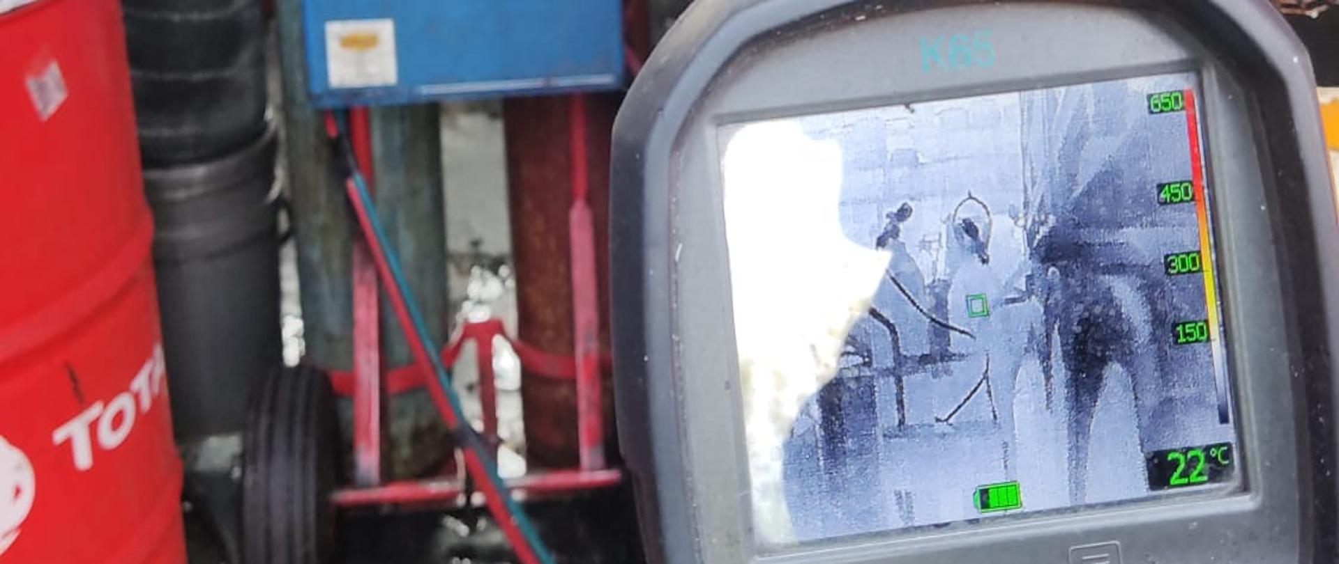 Na zdjęciu na pierwszym planie widać kamerę termowizyjną i rękawicę strażacką. Na ekranie kamery w rysuje się czarnobiały obraz przedstawiający butle gazowe oraz zmierzoną temperaturę wynoszącą 22 st. C. W tle widoczne dwie butle gazowe - jedna koloru niebieskiego z tlenem, a druga koloru kasztanowego z acetylenem. Butla z acetylenem nosi czarne po pożarowe ślady. Tuż obok butli stoi samochód ciężarowy.