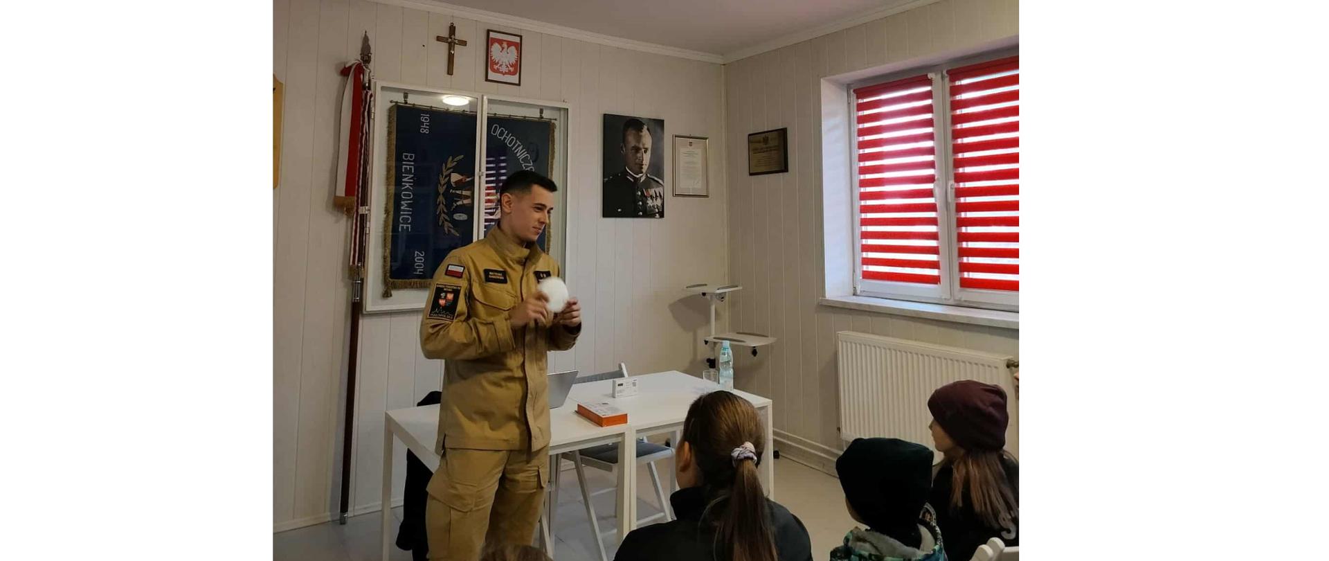 Strażak trzyma w ręce czujkę, którą prezentuje grupie młodych ludzi. Osoby te siedzą na krzesłach. W tle widać zawieszoną szafkę ze sztandarem oraz godło Polski i portret rtm. Witolda Pileckiego.