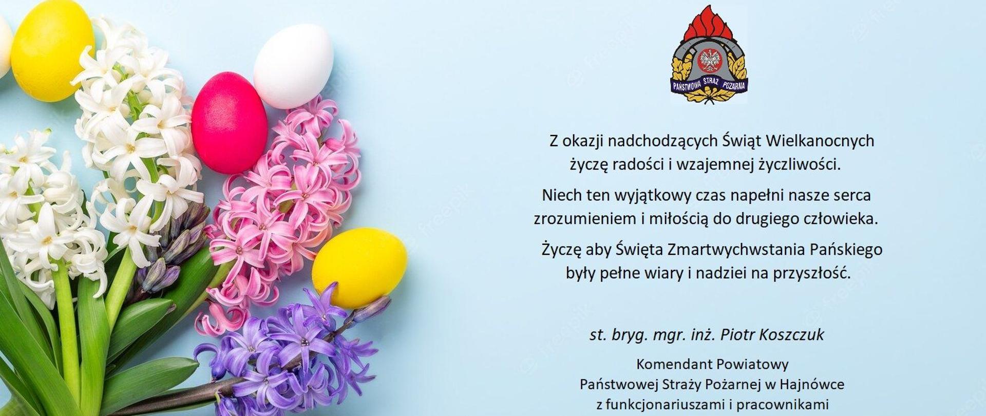 Na jasnoniebieskim tle z lewej strony bukiet kwiatów z kraszankami. Po prawej stronie u góry logo PSP oraz życzenia z okazji świąt Wielkanocnych Komendanta Powiatowego PSP w Hajnówce.