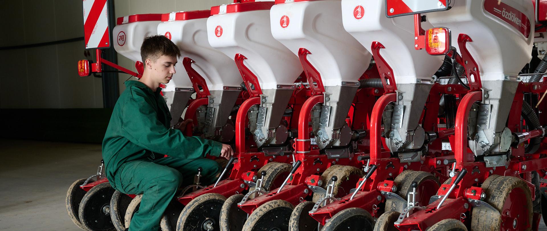 Uczeń w zielonym stroju roboczym siedzi z lewej strony przy siewniku i wykonuje czynności regulacyjne. 