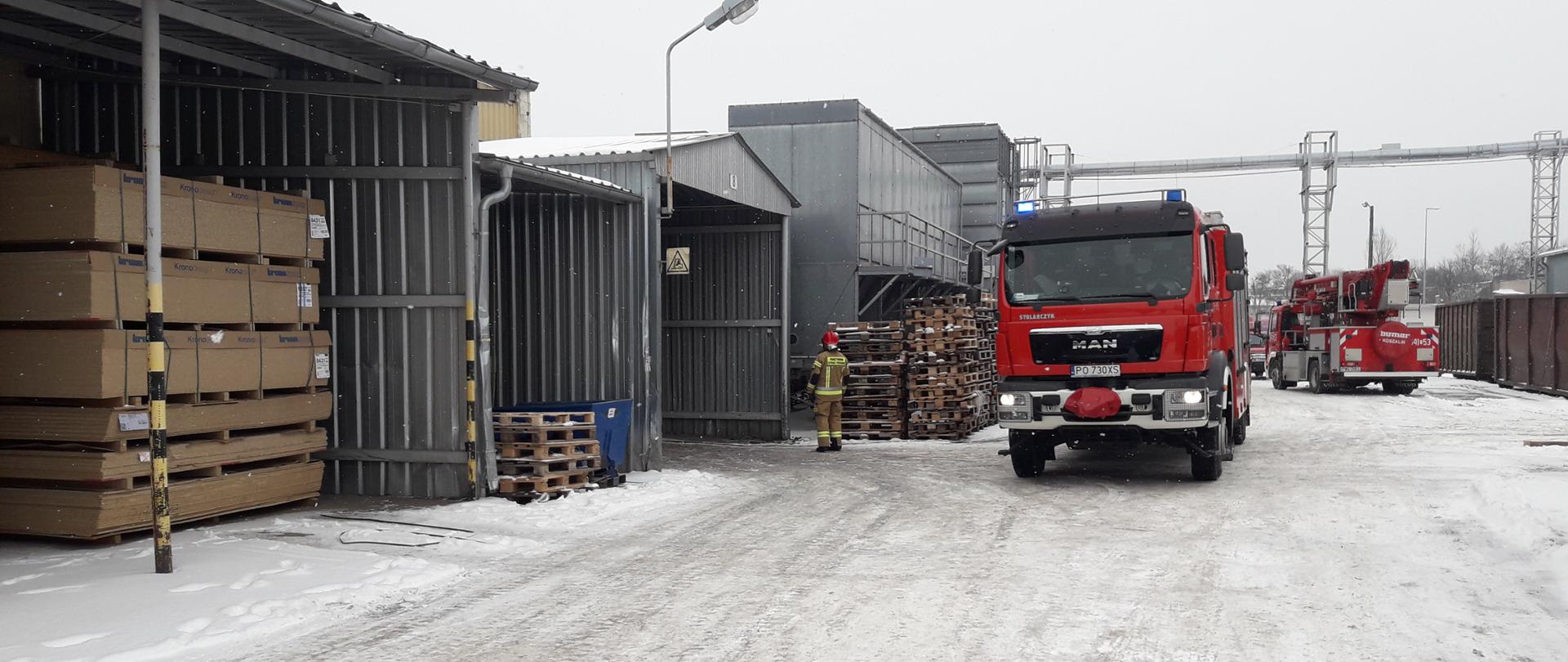 Plac na terenie zakładu. Na środku samochód pożarniczy. Na drugim planie strażak oraz podnośnik hydrauliczny.