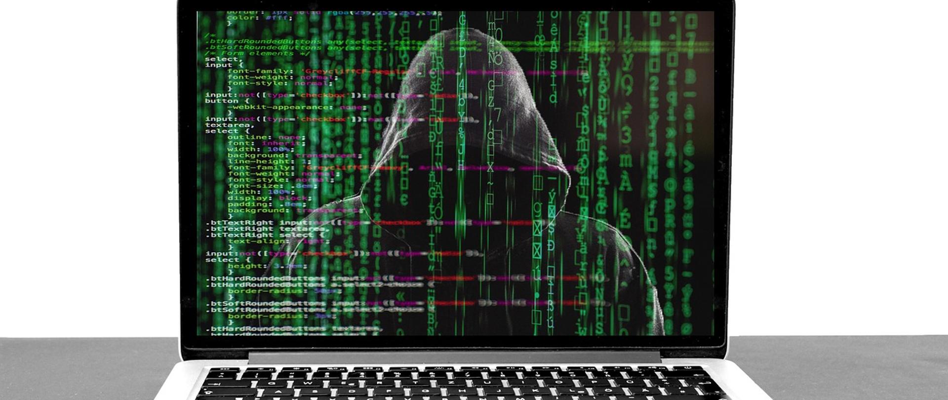 laptop, na ekranie laptopa widać postać w kapturze i dużo zielonych cyferek i literek porozrzucanych na całym ekranie