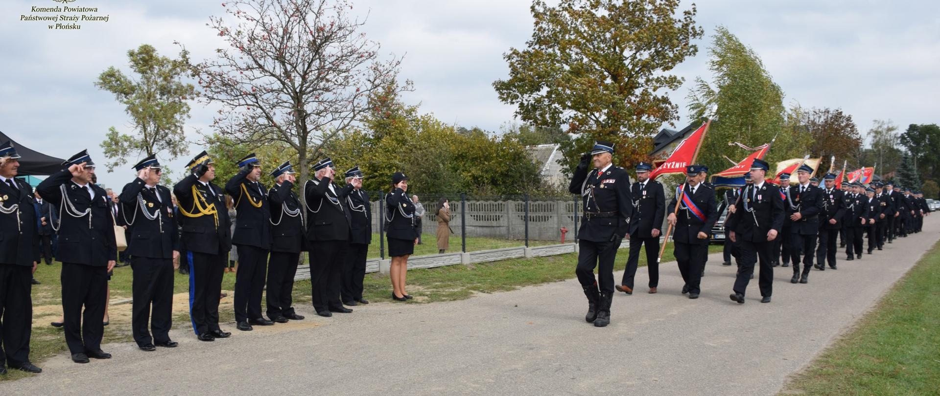 Drogą maszeruje kolumna strażaków, z lewej strony goście honorowi przyjmujący defiladę.
