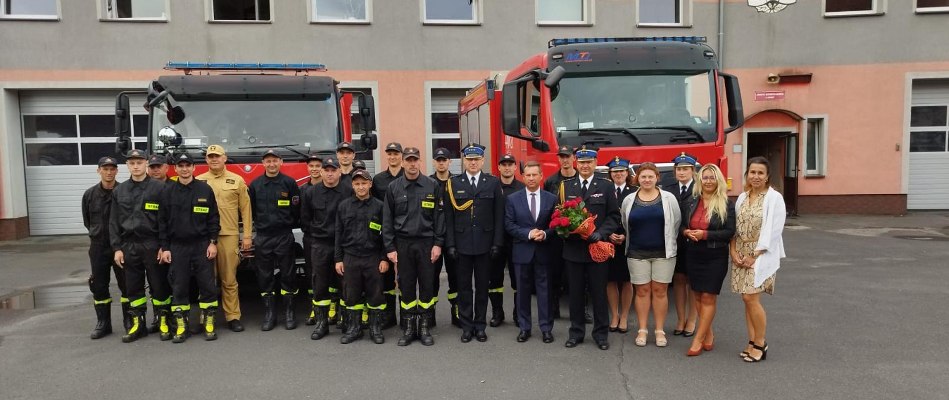 Obraz przedstawia strażaków pozujących do wspólnego zdjęcia. W tle samochody pożarnicze i budynek komendy. Pora dzienna.