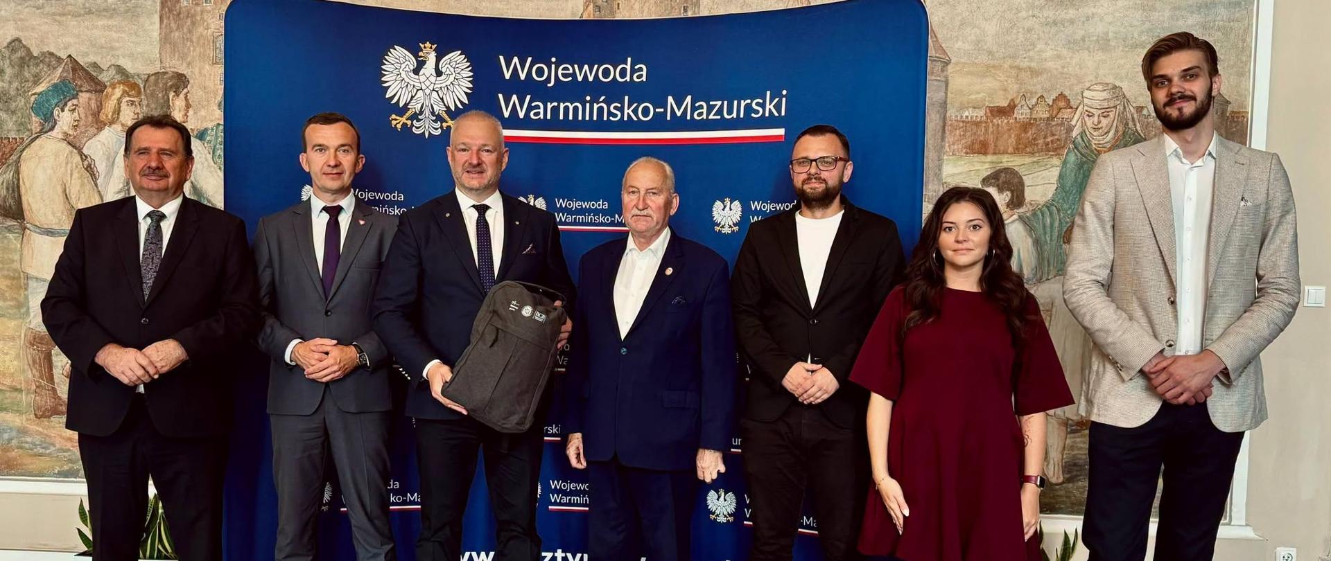 Grupa mężczyzn w garniturach w raz z kobietą w sukience stoi na tle granatowej ścinki, jeden mężczyzna trzyma w rękach plecak 