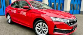 Czerwony samochód marki Skoda. Biały napis Straż oraz boczny numer operacyjny znajdujący się na boku pojazdu. Stoi na kostce pol brukowej a w tle niebieskie bramy garażowe budynku KM PSP w Gdyni.