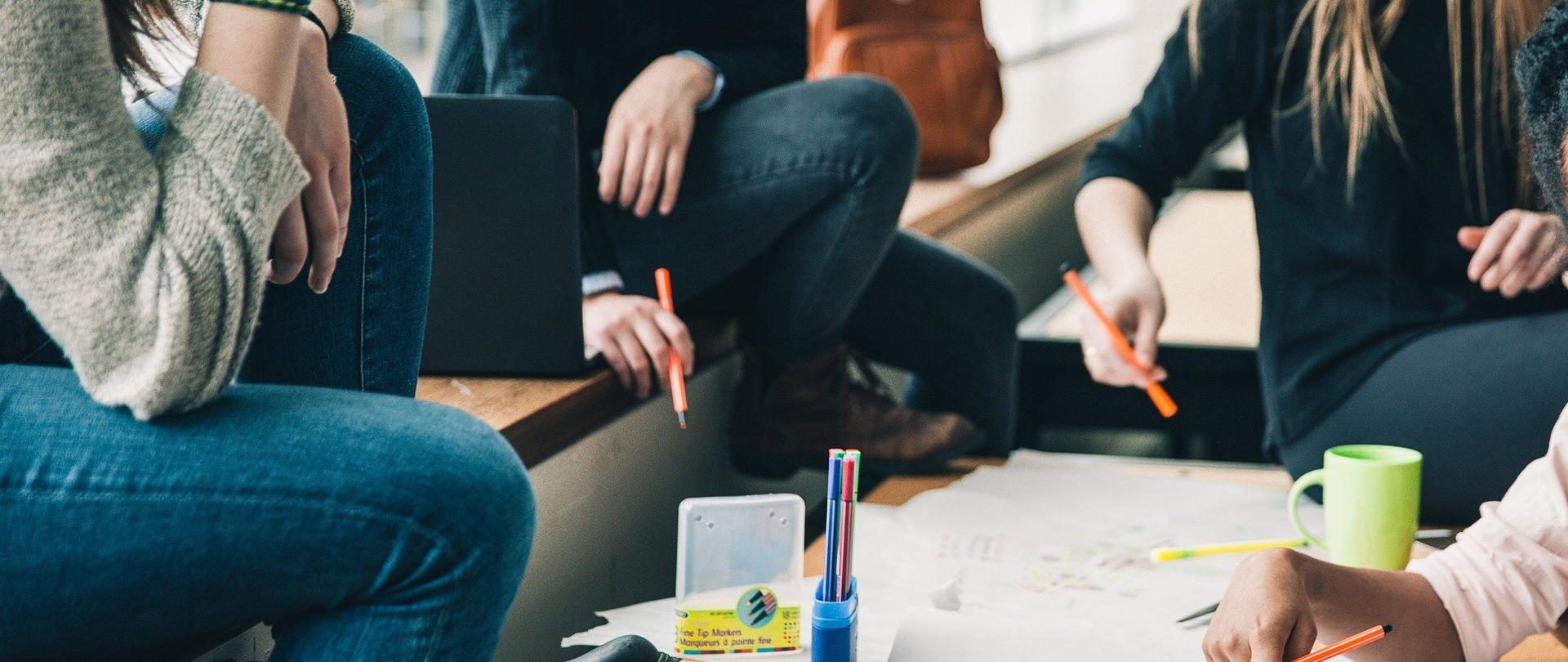 Grafika przedstawia sylwetki młodych osób, które siedzą wspólnie nad planem narysowanym na papierze.
