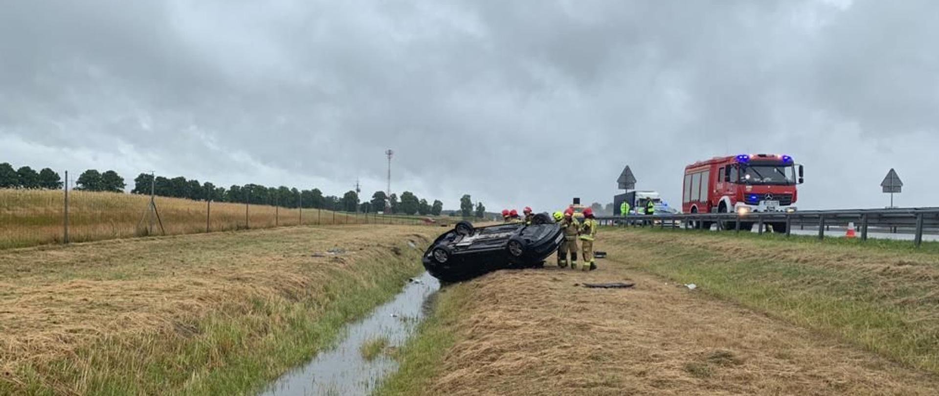 Dzień, opady deszczu. Samochód osobowy leżący na dachu w rowie poza jezdnią autostrady A2.