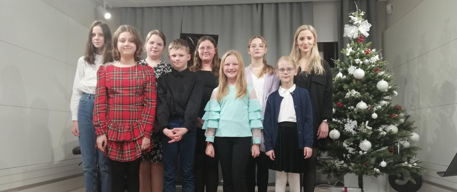 Zdjęcie przedstawia uczniów wraz z pedagogiem stojących w dwurzędzie na scenie. Z prawej strony stoi ubrana choinka a pod nią prezenty zawinięte w szary i czerwony papier z czerwonymi wstążkami. W tle widać zasłonięte kotary w szarym kolorze. 
