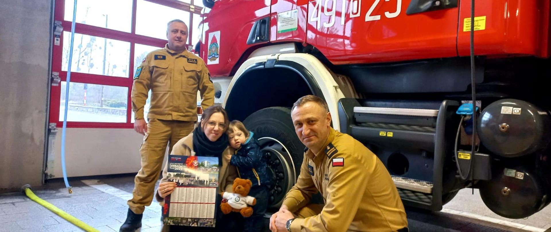Na zdjęciu znajduje się samochód pożarniczy dwóch strażaków, mama oraz dziecko. 