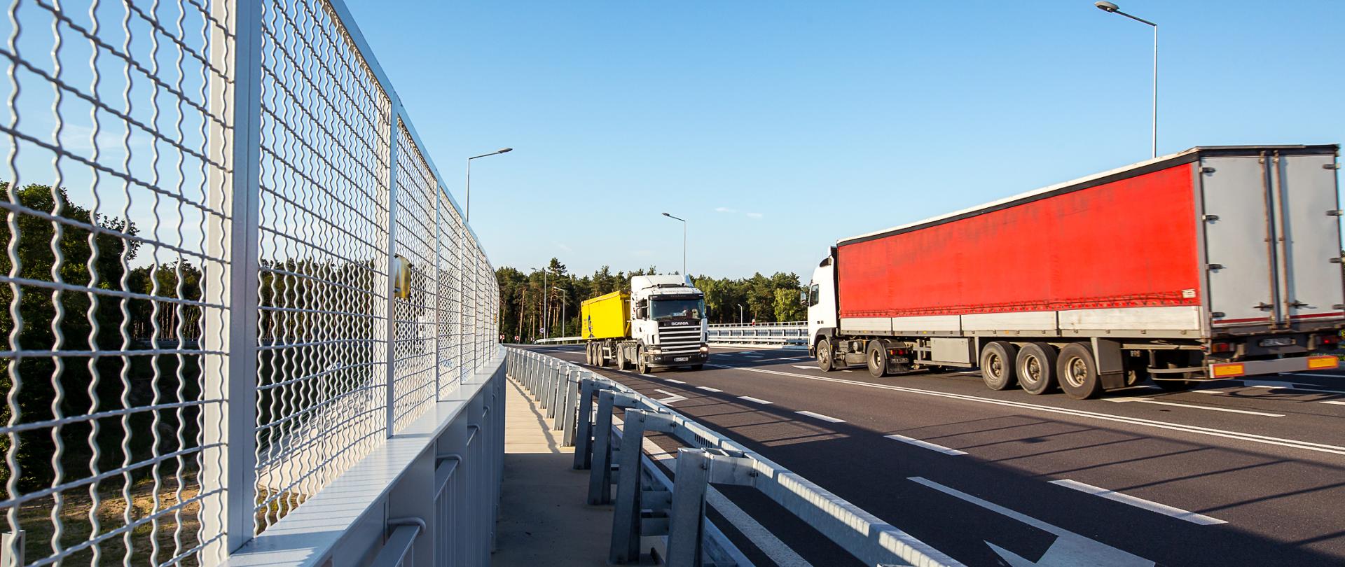 Świeci słońce. Widok z poziomu ziemi na drogę, po której jadą ciężarówki. Po obu stronach widać bariery. 