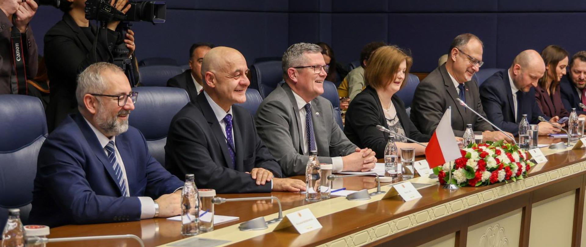 Delegation members – five women and two men – are seated at a conference table during bilateral talks. All are dressed in formal business attire: men in suits and ties, women in blazers or elegant blouses.