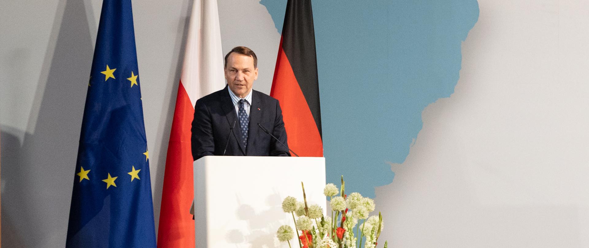 Deputy Prime Minister Sikorski wearing a navy suit and blue tie speaks from a lectern during the Polish-German Forum in Berlin. Behind him are the flags of the European Union, Poland, and Germany, as well as a large world map backdrop displaying the text “Deutsch-Polnisches Forum / Polsko-Niemieckie Forum, Berlin, 17.06.2026.” On the right side of the stage, a sign language interpreter dressed in dark clothing translates the speech. A large audience is seated in rows facing the stage, with some attendees taking photos of the event using their phones.