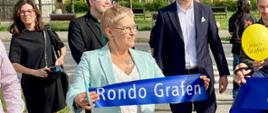 Wiceminister Mrówczyńska stoi na wyłożonym szarą kostką chodniku, trzyma kawałek granatowej wstęgi z napisem rondo Grafen.