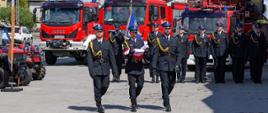 Poczet flagowy strażaków PSP niesie flagę Polski podczas oficjalnego apelu na tle wozów bojowych.