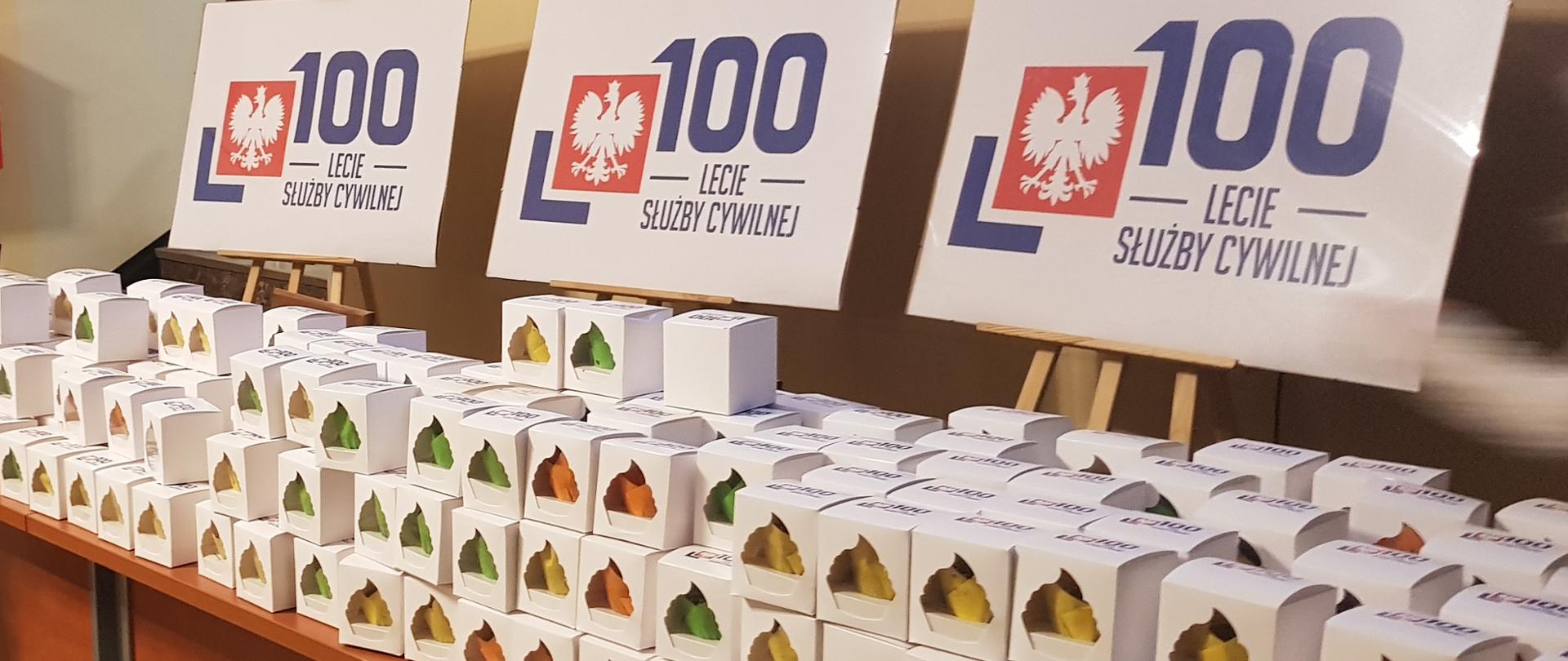 Na długim stole pudełka z upominkami dla pracowników Zachodniopomorskiego Urzędu Wojewódzkiego w Szczecinie z okazji 100-lecia służby cywilnej. Ponad stołem tablice z symbolem obchodów tego jubileuszu.