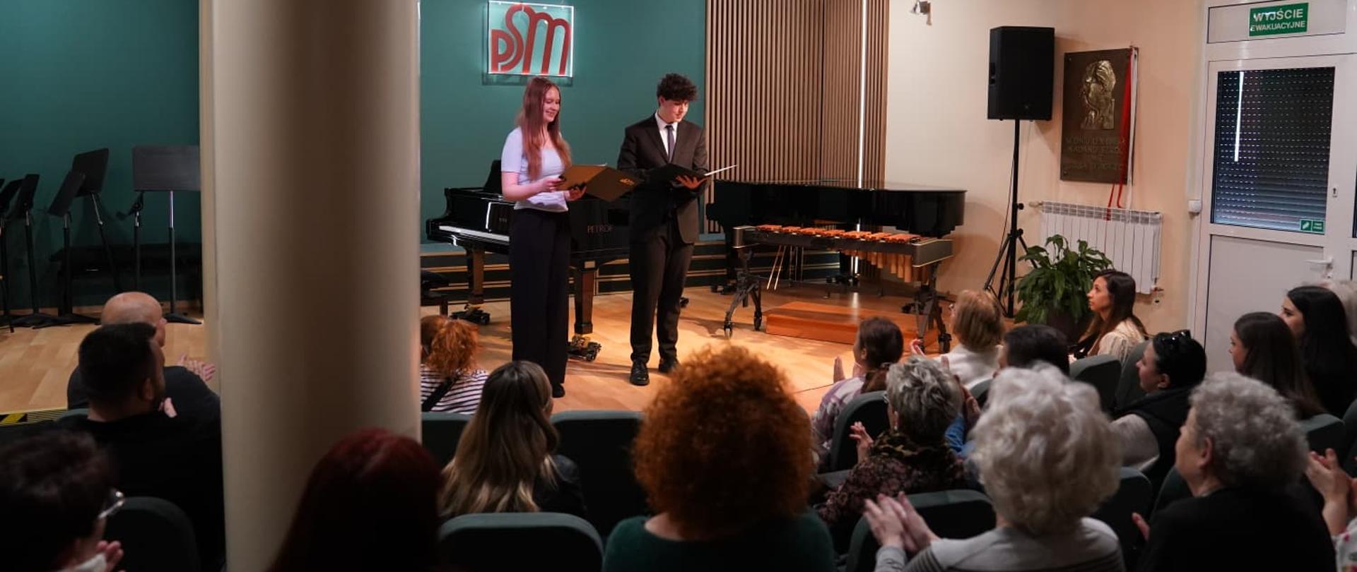 Na zdjęciu widać publiczność siedzącą i oklaskującą dwoje prowadzących koncert uczniów