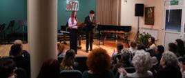 Na zdjęciu widać publiczność siedzącą i oklaskującą dwoje prowadzących koncert uczniów
