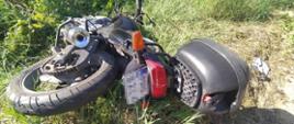 Przydrożny rów porośnięty trawą . W nim leżący motocykl biorący udział w wypadku. Jednoślad koloru czarnego, widoczny od tyłu.