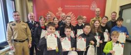 Dzieci z przodu, dorśli z tyłu stoją na tle ścianki firmowej w kolorze czerownym z napisem Państwowa Straż Pożarna i logo PSP. Dzieci trzymają w rękach dyplomy i upominki.