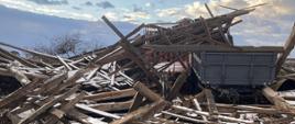 Na środku zdjęcia dwie przyczepy zasypane stertą desek, dookoła leżą porozrzucane deski przez silne wiatry, niebo ciemne, burzowe z miejscowymi przejaśnieniami