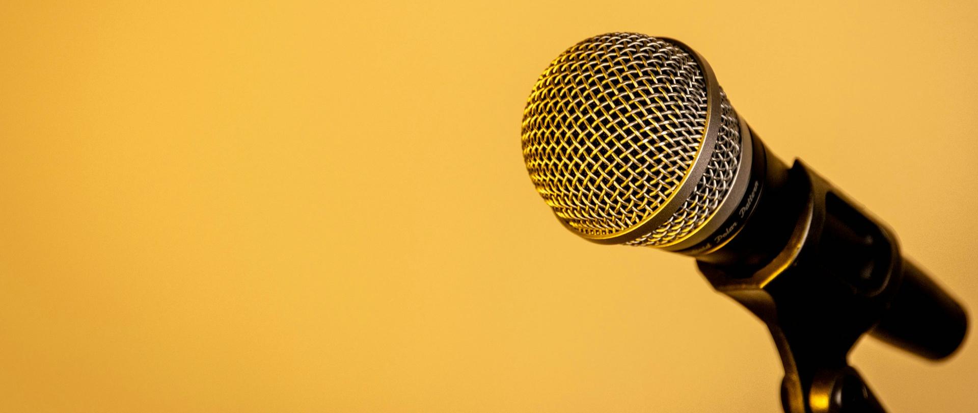 Mikrofon na statywie ustawiony na tle żółtej ściany, przygotowany do wystąpienia lub nagrania głosu.