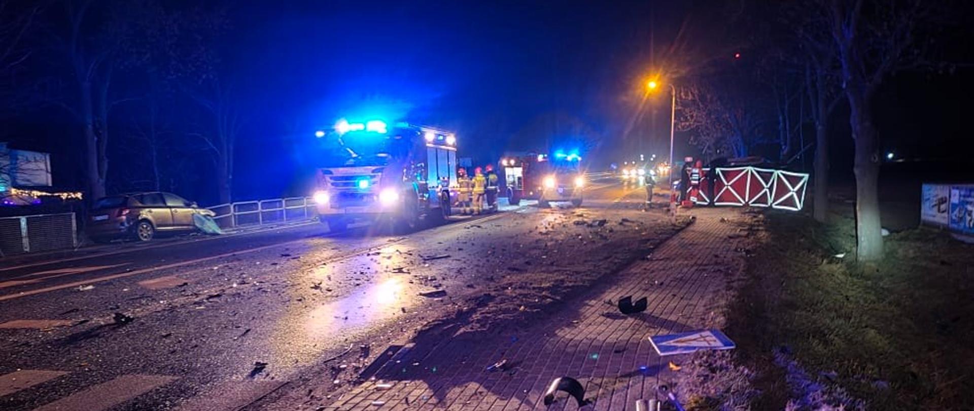 Widok na miejsce zdarzenia, zdjęcie zrobione o zmroku. Widoczne dwa rozbite samochody osobowe stojący po obu stronach jezdni. Na drodze widoczne samochody straży pożarnej z włączonymi sygnałami błyskowymi. Przy jednym z aut rozstawiony czerwony parawan.