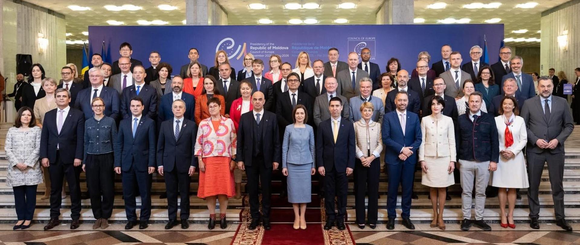 The photo shows a group of about 20 people. They stand and look towards the lens. Behind them is a blue wall with the inscription "Republic of Moldova".