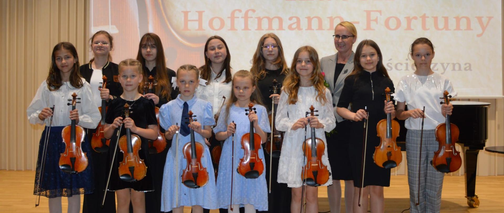 Zdjęcie zostało wykonane podczas koncertu klasy skrzypiec Pani Izabeli Hoffmann- Fortuny. Na scenie stoi grupa odświętnie ubranych dziewczyn. Wszystkie trzymają skrzypce. Wśród nich kobieta blondynka w okularach w czarnej sukience i szarej marynarce. W tle slajd z napisem "koncert klasy skrzypiec Izabeli Hoffmann- Fortuny".