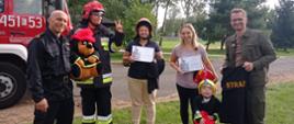 Sześć osób pozuje do fotografii, dwie kobiety trzymają przed sobą pamiątkowe dyplomy uczestników zdjęcia, którzy ubrani są w ubrania bojowe strażaków, dwóch chłopców i jedna kobieta maja na głowie hełm strażacki, druga kobieta trzyma pod pachą hełm strażacki, mężczyzna obok niej trzyma koszulkę barwy czarnej z napisem STRAŻ skierowanym do przodu, po lewej stronie fotografii stoi umundurowany strażak w ubranie barwy czarnej i trzyma pluszową maskotkę ubrana w strażackie ubranko z hełmem w kolorze czerwonym, w tle pojazd ciężarowy straży pożarnej z numerem bocznym 451E53 barwy czerwonej oraz drzewa, wszyscy stoją na zielonej trawie, słońce za chmurami, bezwietrznie