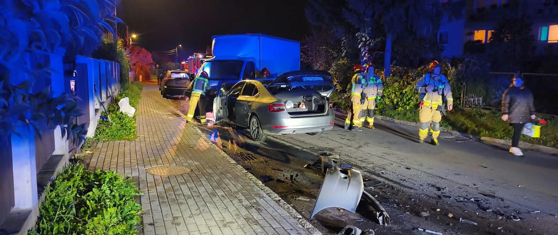 Zdjęcie przedstawia nocne działania służb ratowniczych na ulicy osiedlowej po zdarzeniu drogowym. Na jezdni znajduje się uszkodzony samochód osobowy z otwartymi drzwiami i klapą bagażnika. Wokół pojazdu widoczne są porozrzucane elementy karoserii oraz inne pozostałości po zdarzeniu. Strażacy ubrani w odzież ochronną i hełmy prowadzą działania przy samochodzie. Miejsce zdarzenia oświetlają latarnie uliczne oraz niebieskie światła pojazdów uprzywilejowanych.