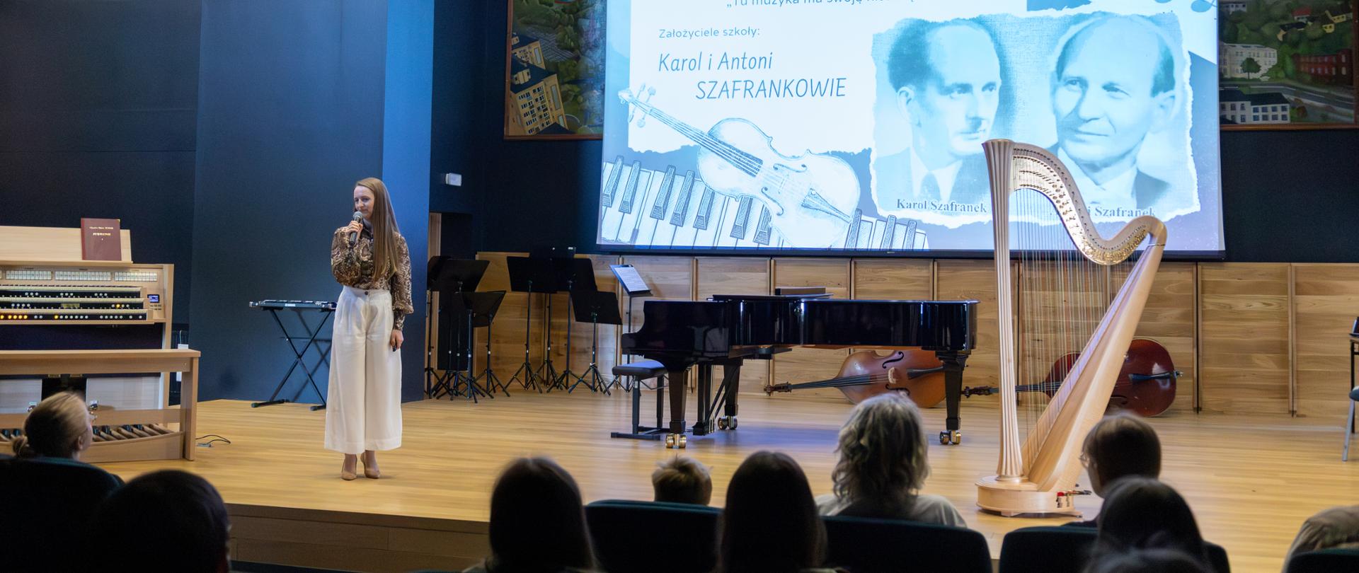 Na scenie stoi kobieta w beżowej koszuli i białych spodniach, prowadząca wykład. Za nią wyświetlana jest prezentacja multimedialna z napisem "MODUŁ I - TRADYCJA oraz założyciele szkoły Karol i Antoni SZAFRANKOWIC" wraz z ich historycznymi fotografiami oraz grafiką przedstawiającą nuty i klawisze fortepianu. Na scenie ustawione są instrumenty muzyczne: fortepian, harfa oraz organy. Widownia zajęta przez publiczność, która obserwuje prezentację.