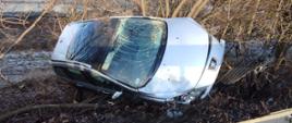 srebrny samochód rozbity poza drogą na skarpie wiaduktu oparty o krzaki oraz drzewa w tle widoczne pola uprawne
