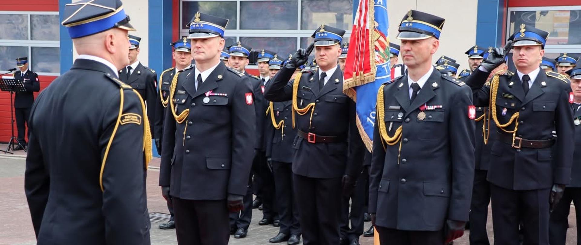 Na placu apelowym dwóch strażaków w mundurze galowym stoją na baczność przed strażakiem przełożonym. W tle poczet sztandarowy ze sztandarem, w dali pododdział strażaków. Widać fragment czerwonego dywanu.