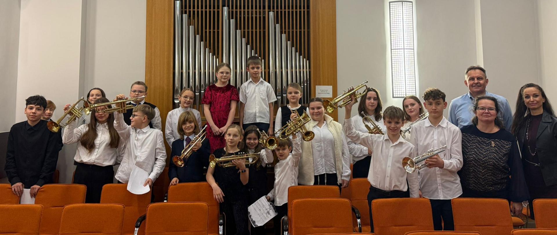 Na zdjęciu widoczna jest grupa uczniów klasy trąbki pozujących w sali koncertowej. Dzieci i młodzież stoją między rzędami pomarańczowych krzeseł, trzymając w rękach trąbki, część z nich unosi instrumenty w górze. Uczniowie ubrani są w stroje koncertowe w jasnych i ciemnych kolorach. W tle znajduje się prospekt organowy z piszczałkami oraz jasne ściany sali. Zdjęcie przedstawia radosny moment po wspólnym występie muzycznym.