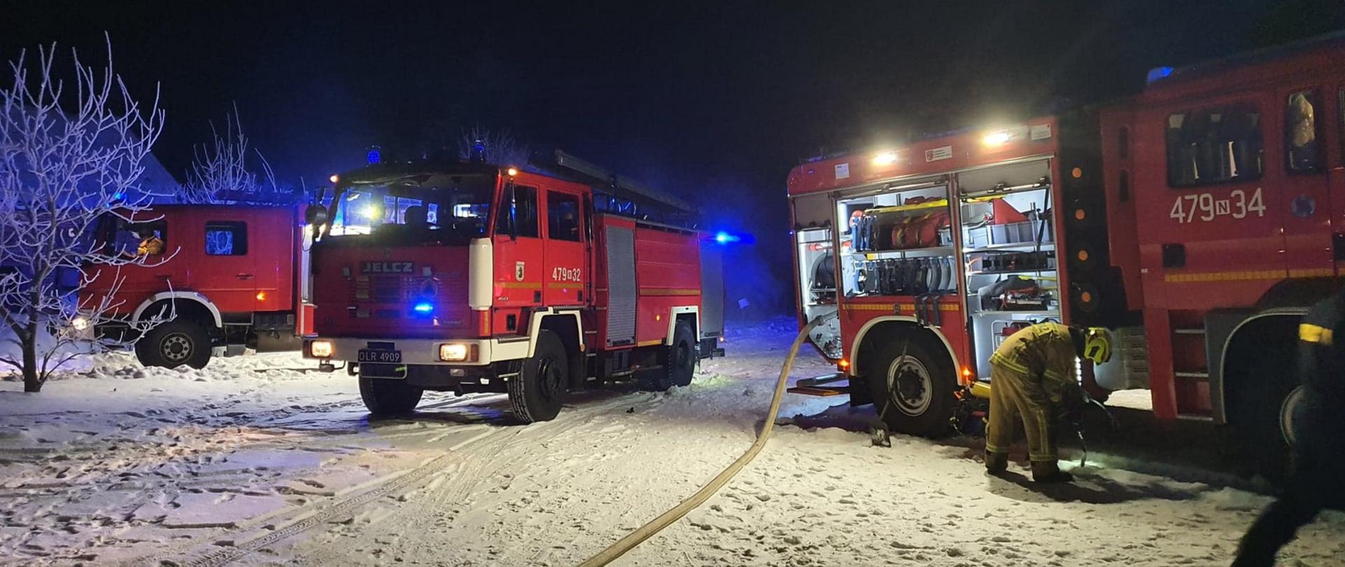 Zdjęcie zrobione w porze nocnej podczas pożaru budynku wielorodzinnego w miejscowości Kępa Tolnicka. Widać trzy czerwone wozy bojowe straży pożarnej stojące na ziemi pokrytej śniegiem, do jednego wozu podłączony jest wąż strażacki, a strażak stojący przy wozie wymienia butlę powietrzną w aparacie Ochrony Układu Oddechowego. 
