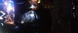 Zdjęcie przedstawia pojazd VW Golf koloru granatowego który uderzył w drzewo. Zdjęcie wykonane nocą. Pojazd widoczny od przodu z uszkodzoną maską silnika i karoserią od strony pasażera. Obok wraku pojazdu postacie strażaków i policjantów
