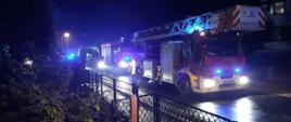 Jest pora nocna. Na zdjęciu widać autodrabinę strażacką, która przyjechały do zdarzenia. Za nim stoi inny samochód strażacki a koło niego karetka pogotowia. Samochody mają włączone niebieskie sygnały błyskowe