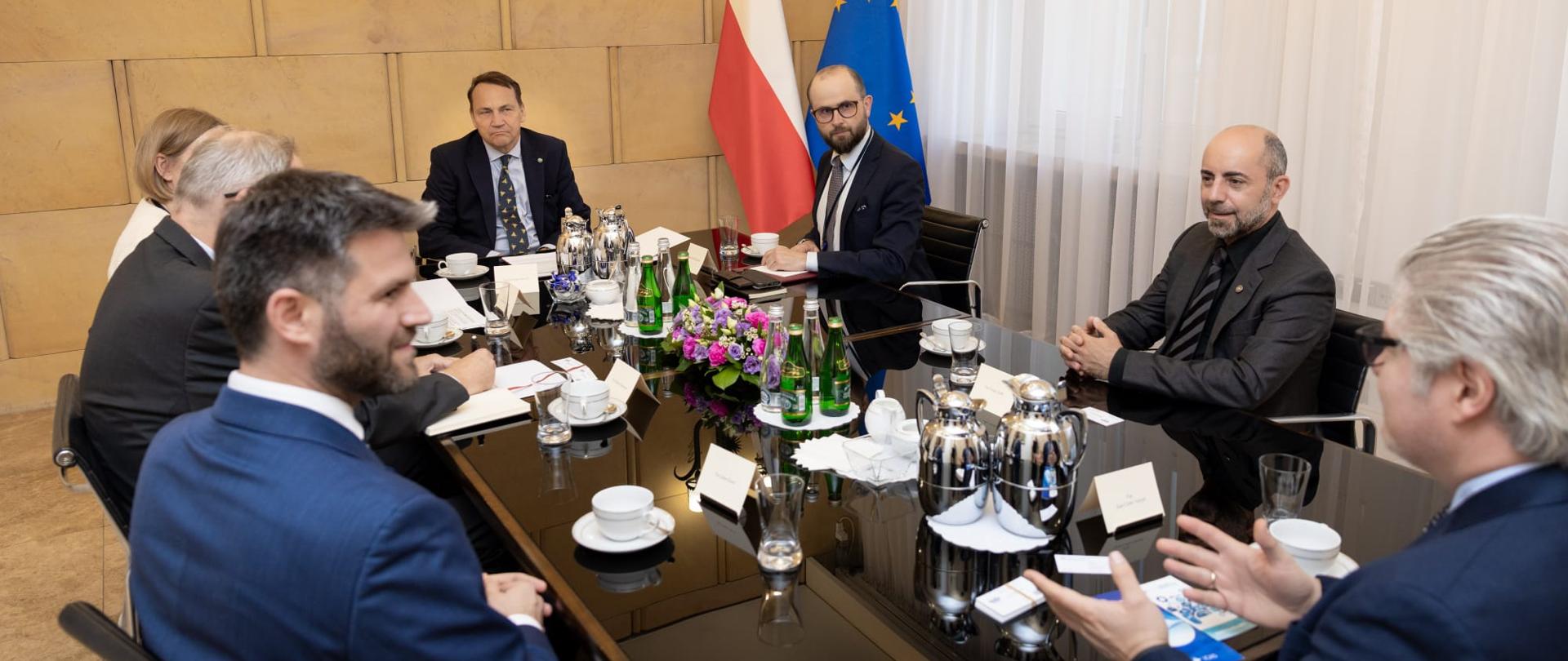 A delegation meeting around a rectangular conference table. On the table are documents, microphones, cups, glasses, water carafes, and an arrangement of pink and purple flowers. The flags of Poland and the European Union are visible in the background.