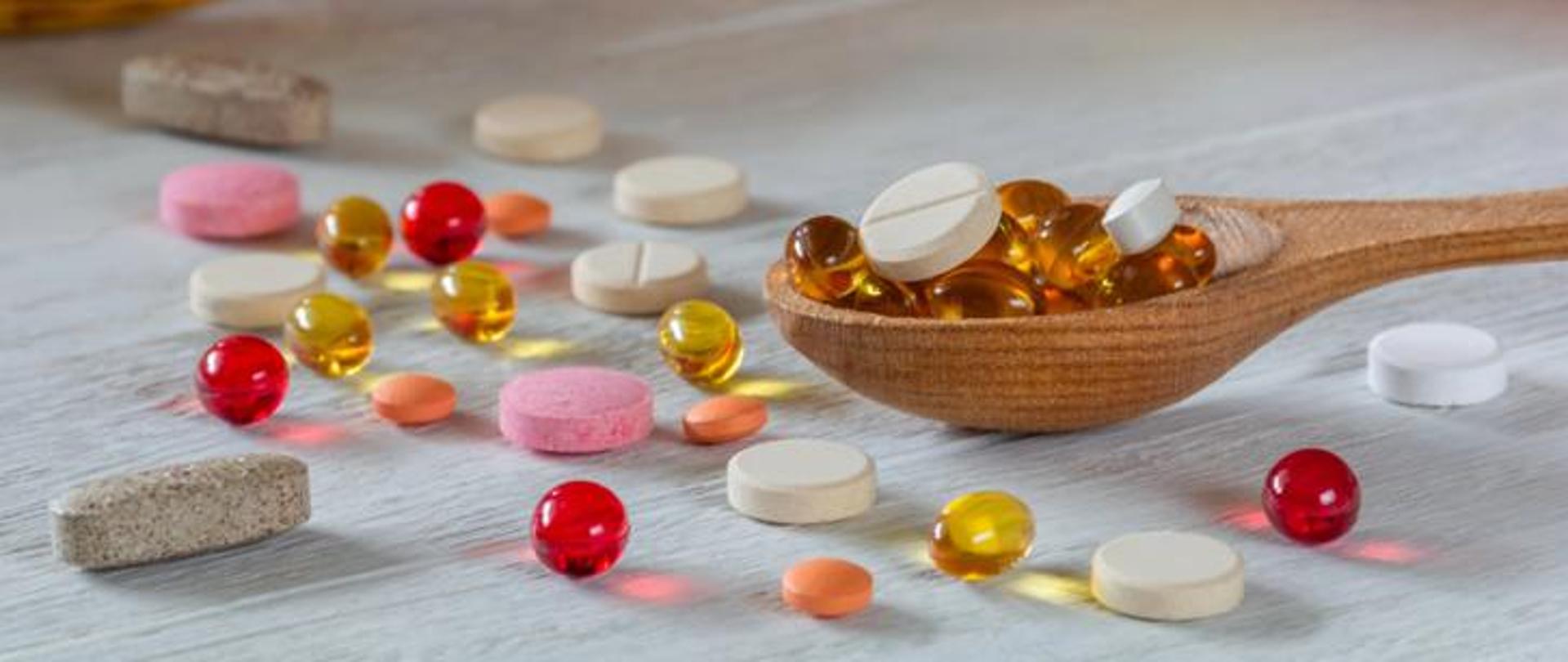 Różnokolorowe tabletki i kapsułki rozrzucone na jasnym drewnianym blacie, część z nich znajduje się w drewnianej łyżce. Wśród suplementów widoczne są tabletki okrągłe, podłużne i żelowe kapsułki w odcieniach czerwieni, żółci i pomarańczu.