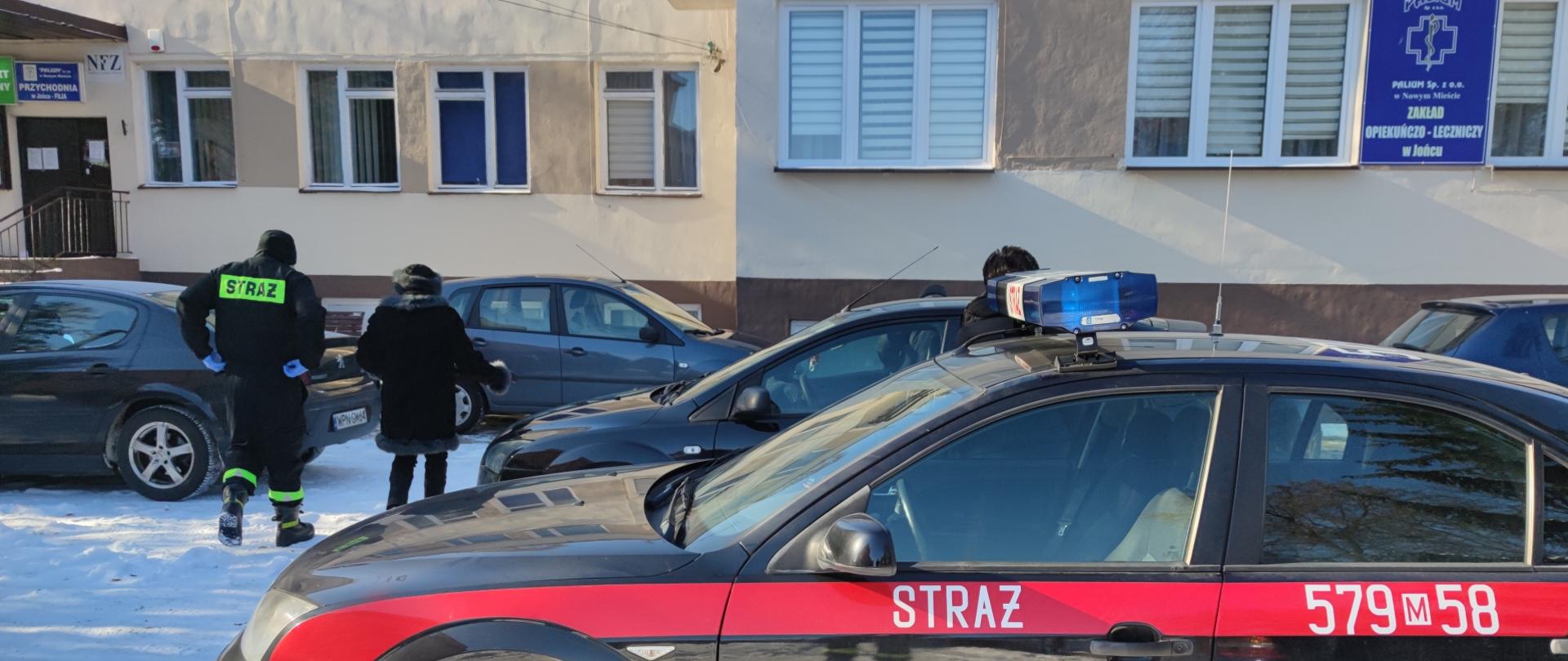 Na pierwszym planie samochód osobowy, oznakowany jako strażacki, w tle budynek - punkt szczepień, z lewej strony osoba udająca się na szczepienie w towarzystwie strażaka OSP. 